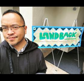 Man with brown skin and short black hair wearing a black hoodie stands in front of a horizontal sign saying "Land Back."